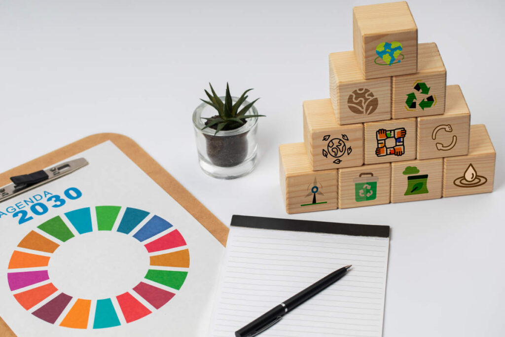 Escritorio con una carpeta con una hoja del logotipo de la Agenda 2030, un cuaderno de notas y unos cubos de madera que representan los ODS